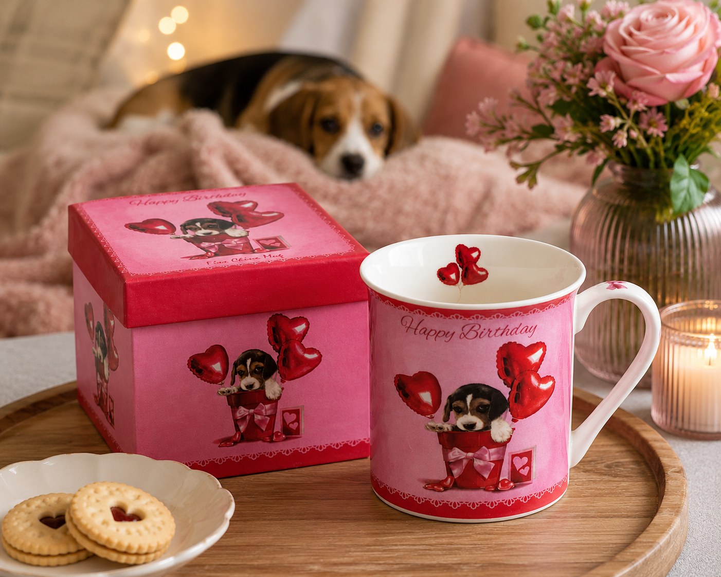 Pink birthday mug with dog design, matching box, cookies, and flowers on a table.