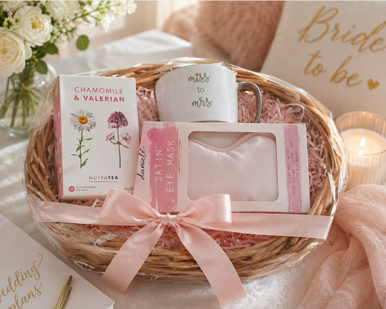 A bridal shower gift basket wrapped in clear cellophane and tied with a pink ribbon, containing a box of chamomile & valerian tea, a white mug that says 'miss to Mrs', a pink packaged satin eye mask, and a pink eye mask, all arranged in a wicker basket with shredded paper. The background is decorated with white roses, a lit candle, and a sign reading 'Bride to be'.